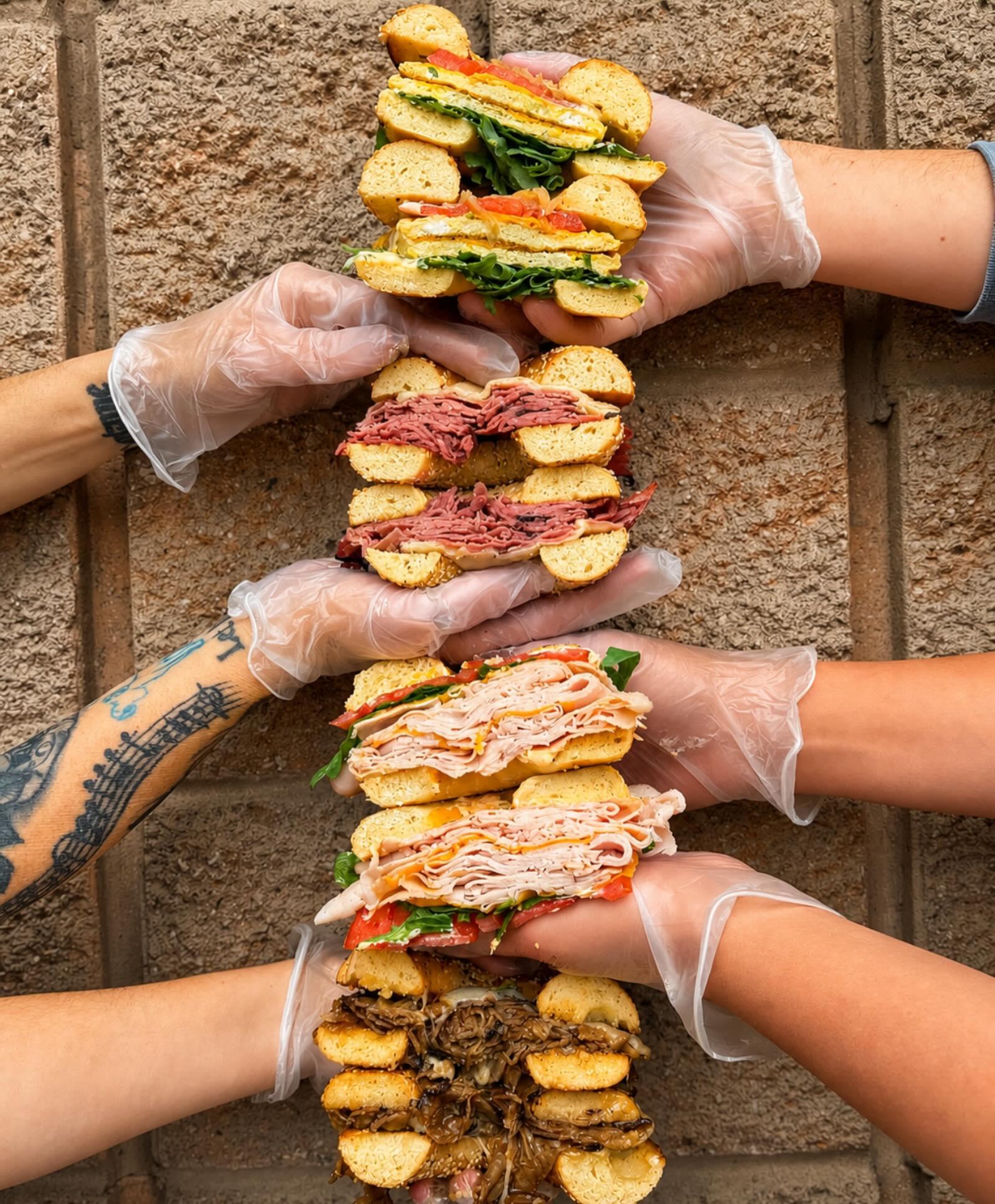 Fresh stacked bagel sandwiches Four pairs of gloved hands holding 4 cut bagel sandwiches